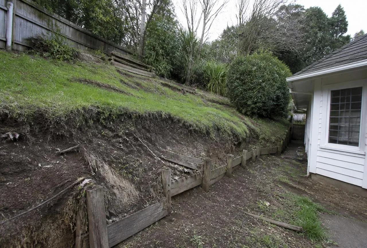 Before: Eroding dirt slope with collapsing timber edging beside a white house before work
